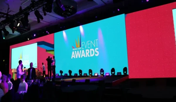 A stage set up for an event with large screens displaying the words "EVENT AWARDS," colorful lighting, and attendees seated at tables.